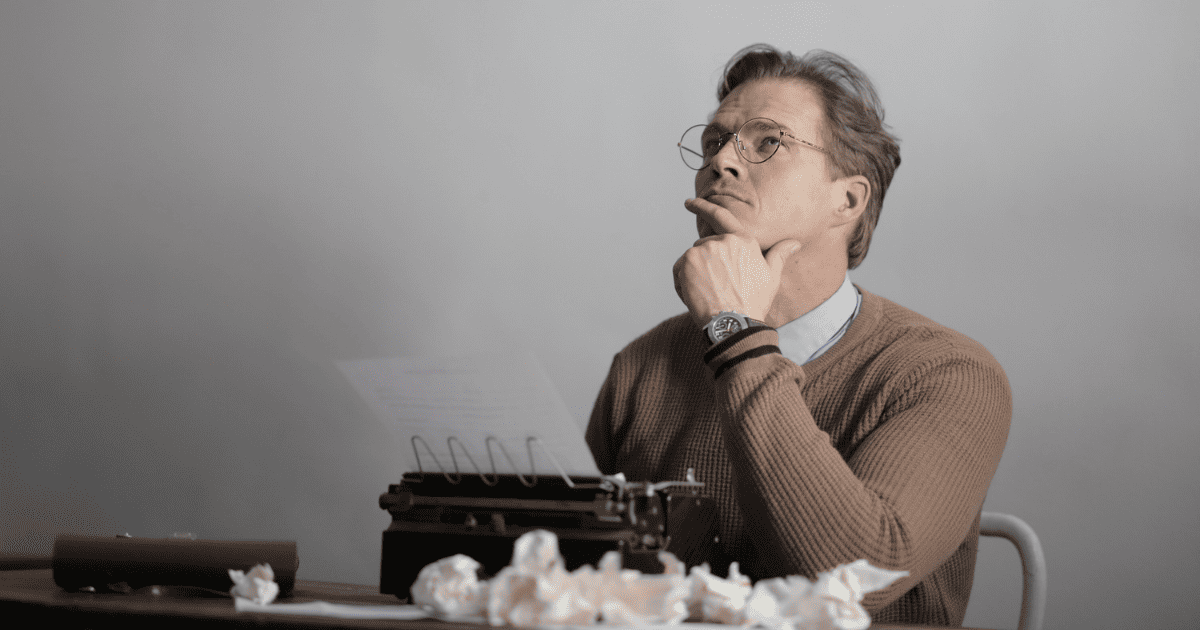 man looking up with hand on chin with typewriter and crumpled paper on table