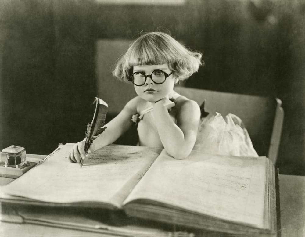 black and white photo of youndg girl with glasses and a large pen writing a book