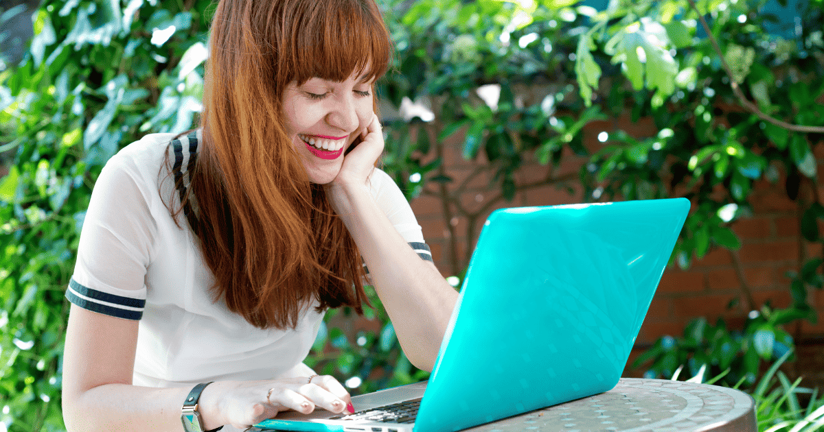 woman smiling while writing outside on blue laptop