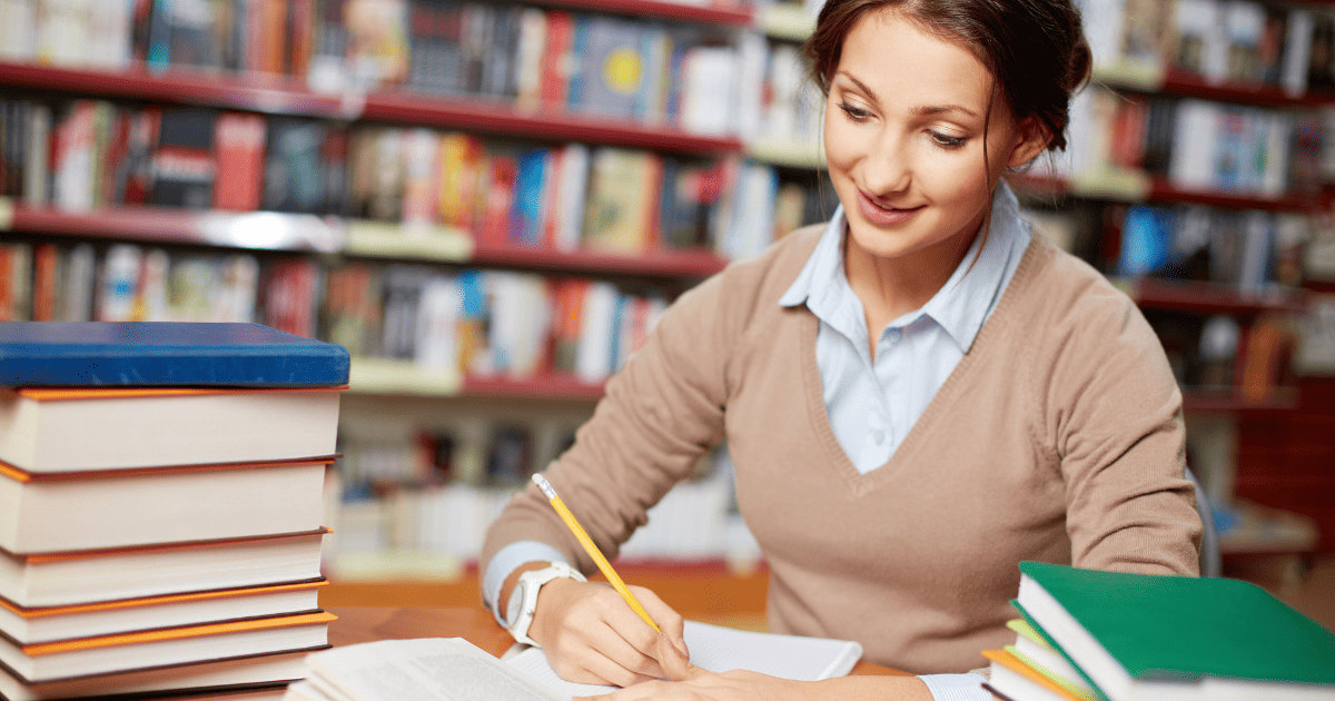 smiling woman writing with pile of books on desk